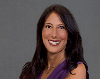 Professional portrait of a smiling woman with long dark hair, wearing a sleeveless purple top, posed in front of a plain gray background.